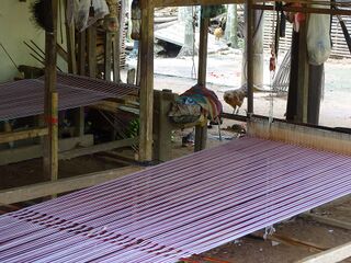 A wooden floor loom under a raised house, with long pale pink warp threads stretched out.