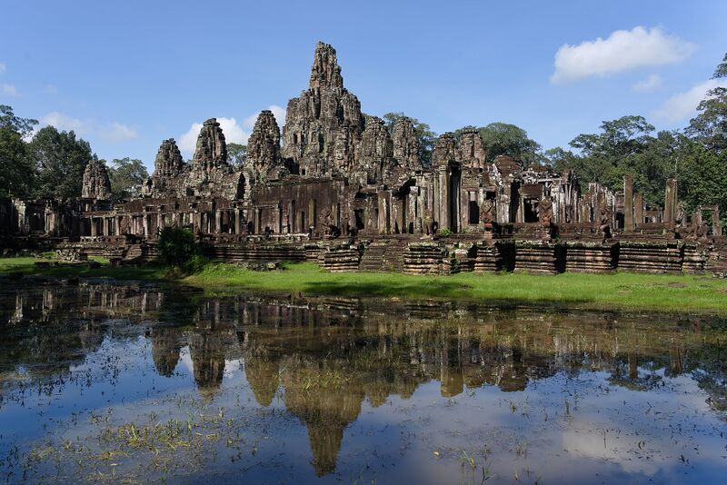 A wide view of the Bayon's crowded towers mirrored in a pond under a blue sky.