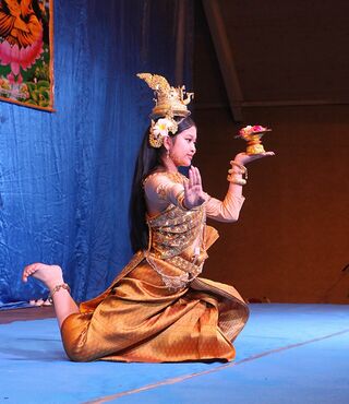 A dancer in a golden costume and tall crown kneels on a blue stage floor, holding a flower offering.
