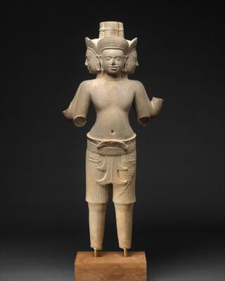 A four-faced sandstone statue in a tall crown, wearing a short sampot pulled up between the legs and folded at the waist.
