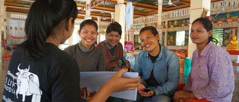 A staff member in a This Life Cambodia T-shirt shows papers to four smiling women seated in a pagoda hall.