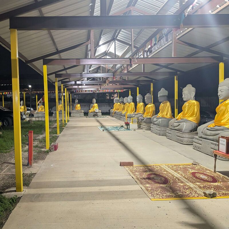 A row of seated stone Buddha statues draped in yellow cloth beside a concrete walkway under a metal roof on yellow posts.