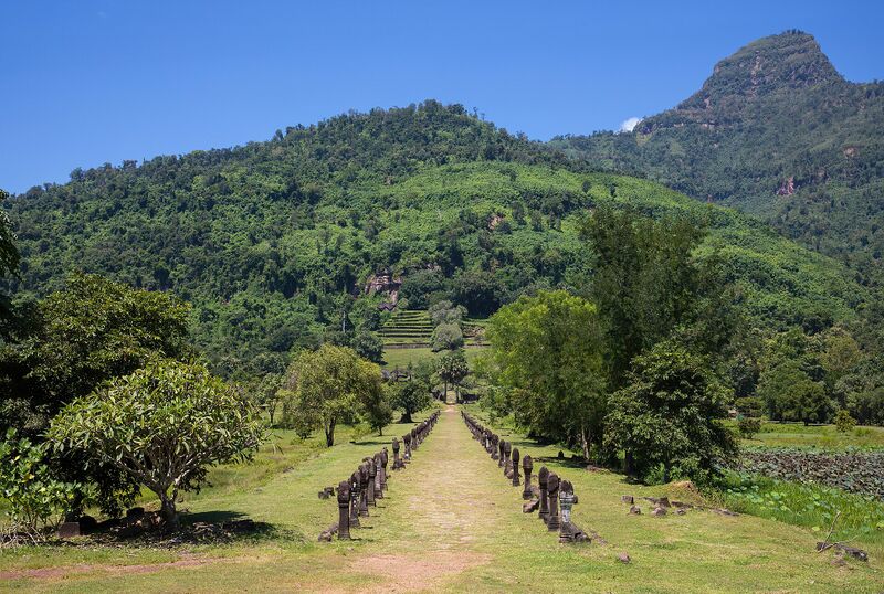 A long straight path lined with short stone posts, leading toward a green mountain with a pointed peak.
