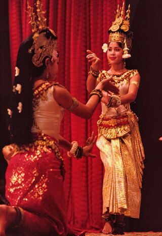 Two dancers in tall gold headdresses and pale costumes posing in front of a red curtain.