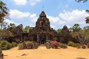 A ruined sandstone temple with a central tower rising over a long gallery wall and two smaller crumbling towers at its sides, behind a sandy courtyard with pink flowering shrubs, scattered carved stones and palm trees, under a blue sky with white clouds.