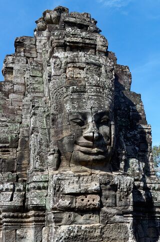 A large serene stone face carved into a weathered tower against a blue sky.