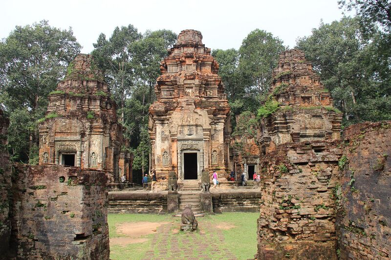 Three weathered brick temple towers with carved doorways on a low platform, seen across a lawn between two ruined brick walls.