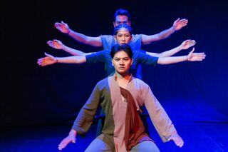 Three dancers stacked one behind another on a blue-lit stage, their arms extended to look like many arms.