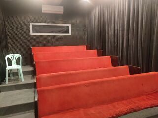 A small room with tiered red benches facing a screen area, dark curtains on the wall and a white plastic chair.