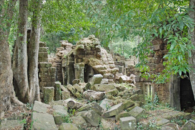 A collapsed stone and brick temple, with fallen blocks and tree roots around it, in a shaded forest.
