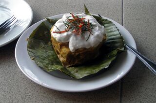 A banana-leaf cup of set fish custard topped with white coconut cream and fine green strips, on a white plate.