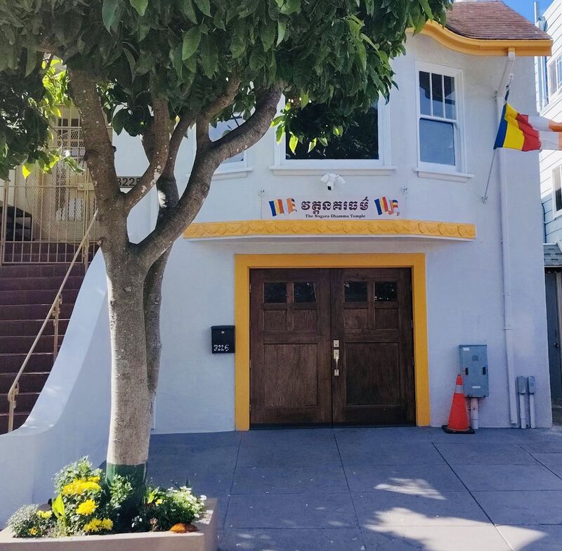 White house front with yellow trim, wooden double doors, a sign in Khmer and English, Buddhist flags and a street tree.