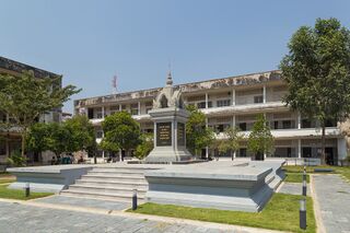 A white memorial with a tiered top on a stepped platform, before three-storey former school buildings.