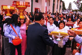 Crowd outside a temple building carrying tall gold offering bowls and trays decorated with red and gold netting.