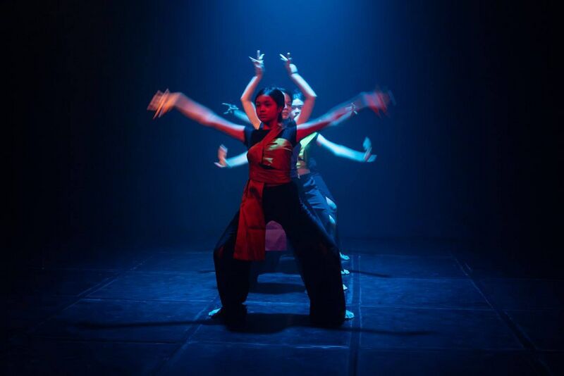 Dancers in red costumes on a dark stage under blue light, arms raised and spread in layered poses.