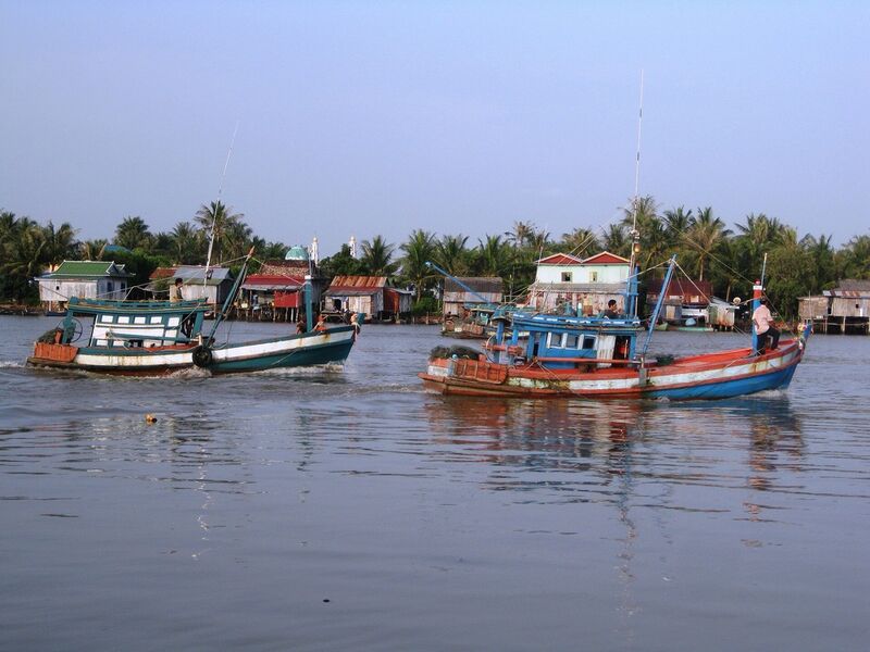 Two fishing boats with blue and red hulls move along a wide river, with stilt houses and palm trees on the far bank.