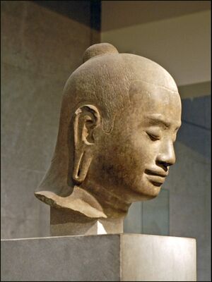 A carved stone head of a man with eyes lowered and a faint smile, his hair drawn up in a small bun, on a museum stand.