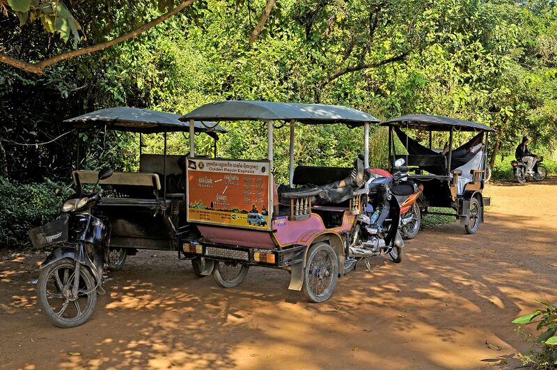 Three canopied tuk-tuks hitched to motorbikes parked in the shade on a dirt lot, one with a route board in Khmer and English, and drivers resting.
