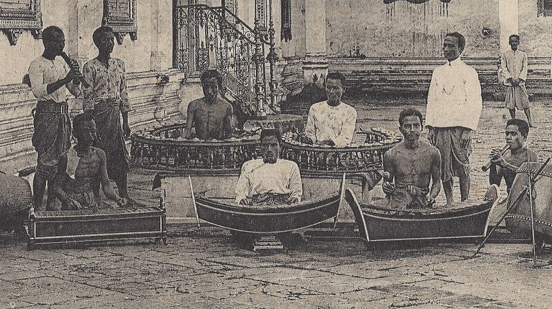 Black-and-white photograph of Cambodian musicians in a palace courtyard, seated at boat-shaped xylophones and circular gong frames, with two men playing reed pipes.