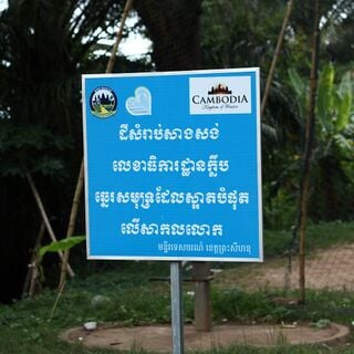 A blue roadside sign with white Khmer script and two small logos at the top, set among palm trees