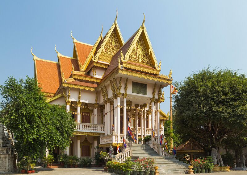 A white Khmer temple hall with several tiered orange and gold roofs behind trees and a flight of steps.