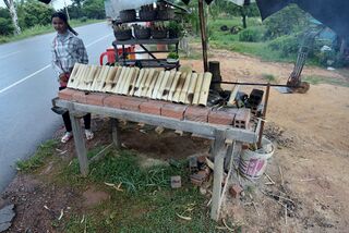 A wooden stand by a road with rows of bamboo tubes on a long grill rack and a vendor standing nearby.