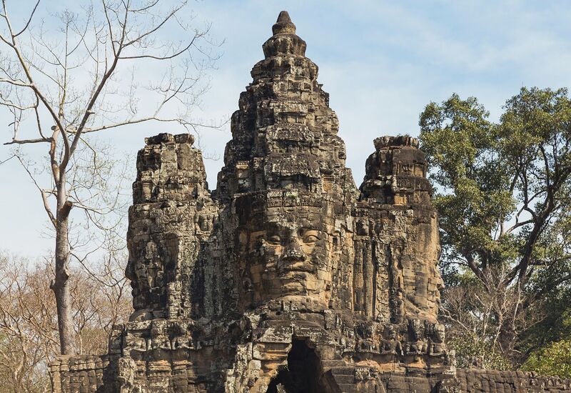 A tall stone gate tower carved with a large serene face, with trees on either side and a pale blue sky.