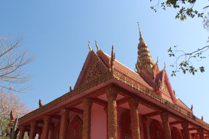 A red Khmer hall with a steep tiered roof and a pointed spire under a blue sky.