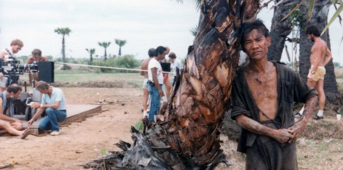 Haing S. Ngor, shirtless and muddy, stands by a palm tree while a film crew works behind him.