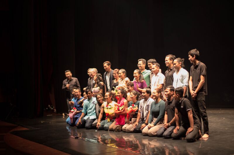 About thirty people gather on a dark stage, some standing and some kneeling in front, smiling and holding bouquets of red, yellow and orange flowers, while a man at the left speaks into a microphone.