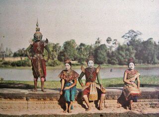 An early color photograph of costumed dancers posing on a stone terrace beside water, trees behind.