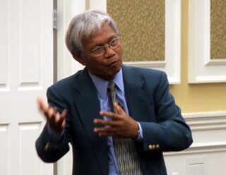 A grey-haired man in a dark suit gestures with both hands as he talks.