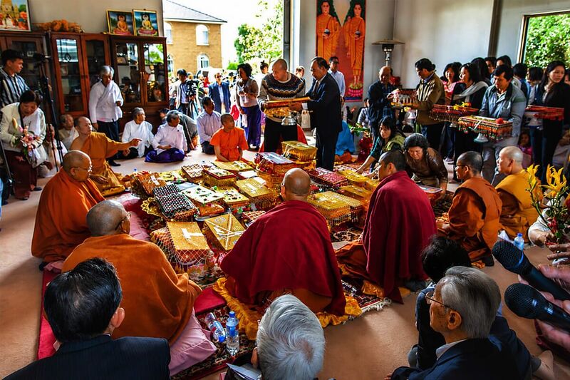 Monks seated around decorated offering baskets on a carpeted floor while lay people stand and kneel around them holding gifts.