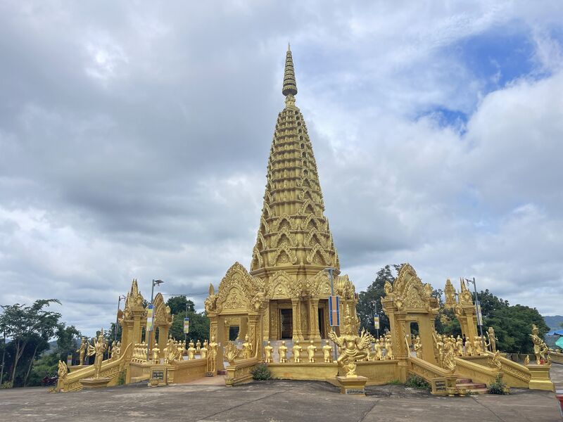A tall gilded tower rises in tiers above a gold shrine with carved gateways on each side. Rows of gold statues line the steps and terraces around it, under a cloudy sky.