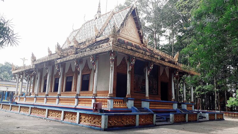 A long Khmer temple hall with a tiered, gilded roof and blue and gold railings, set among tall trees.