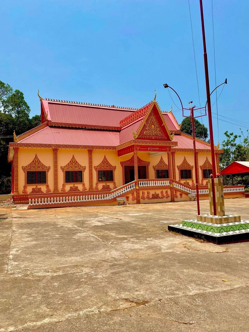A Khmer temple hall with a red tiered roof and ornate orange walls on a wide paved yard beside a flagpole.