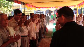 Rows of people with hands pressed together in prayer stand under a decorated tent canopy.