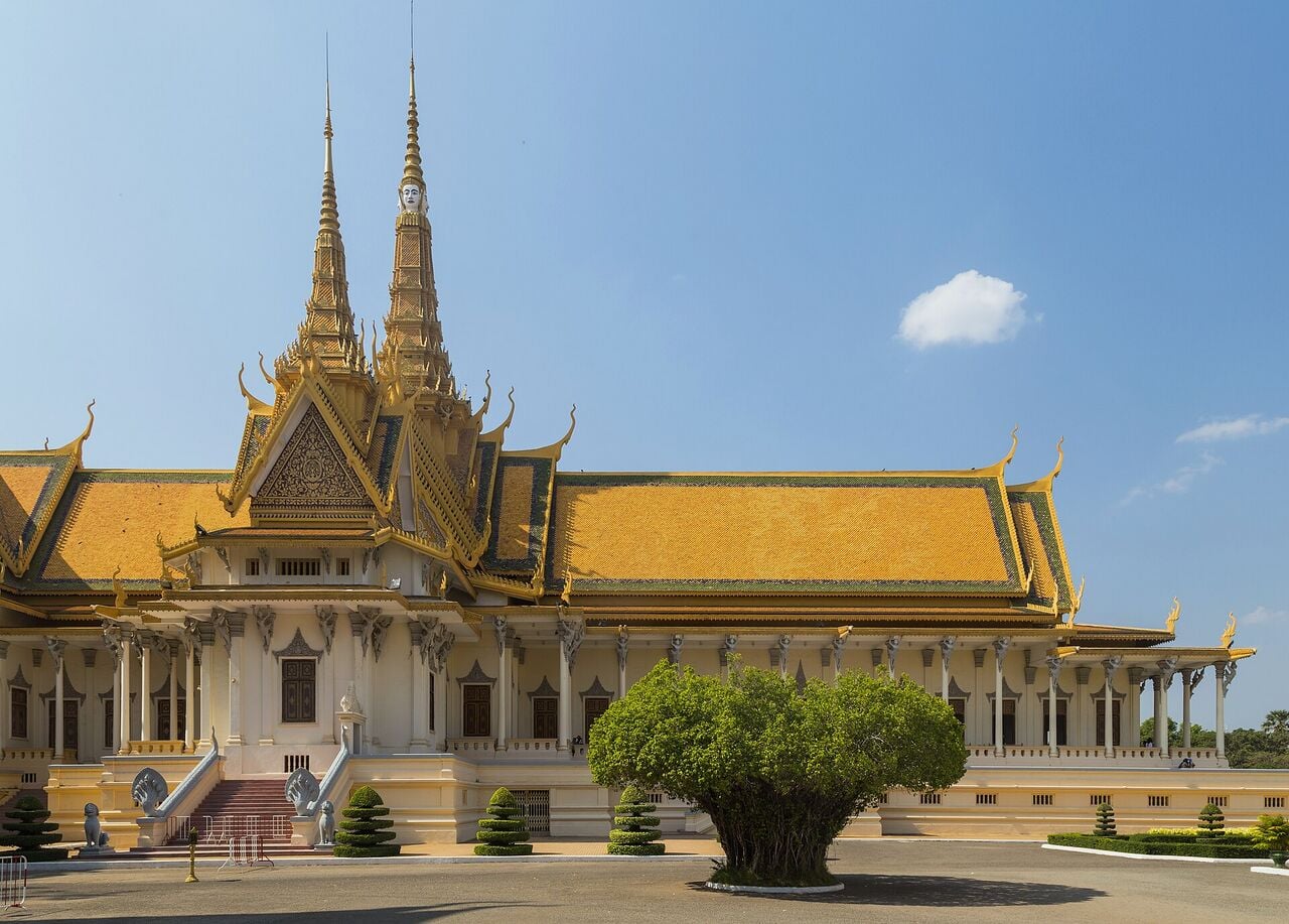 A cream and gold palace hall with a tiered orange roof and tall spires under a blue sky, seen across an empty paved courtyard