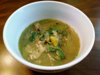 A white bowl of thick green soup with vegetables and meat on a wooden table.