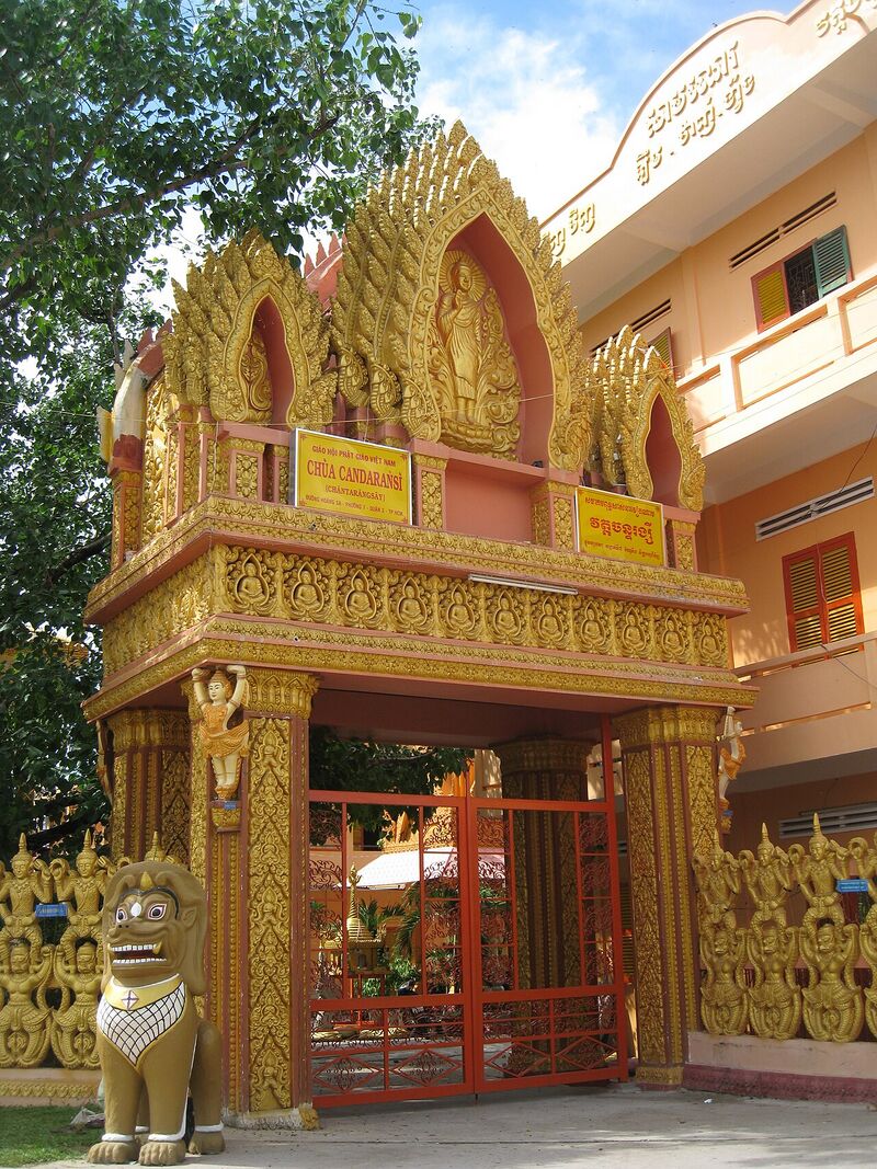 An ornate gilded Khmer-style gateway with red iron gates and a lion statue, beside a pale multi-storey building.