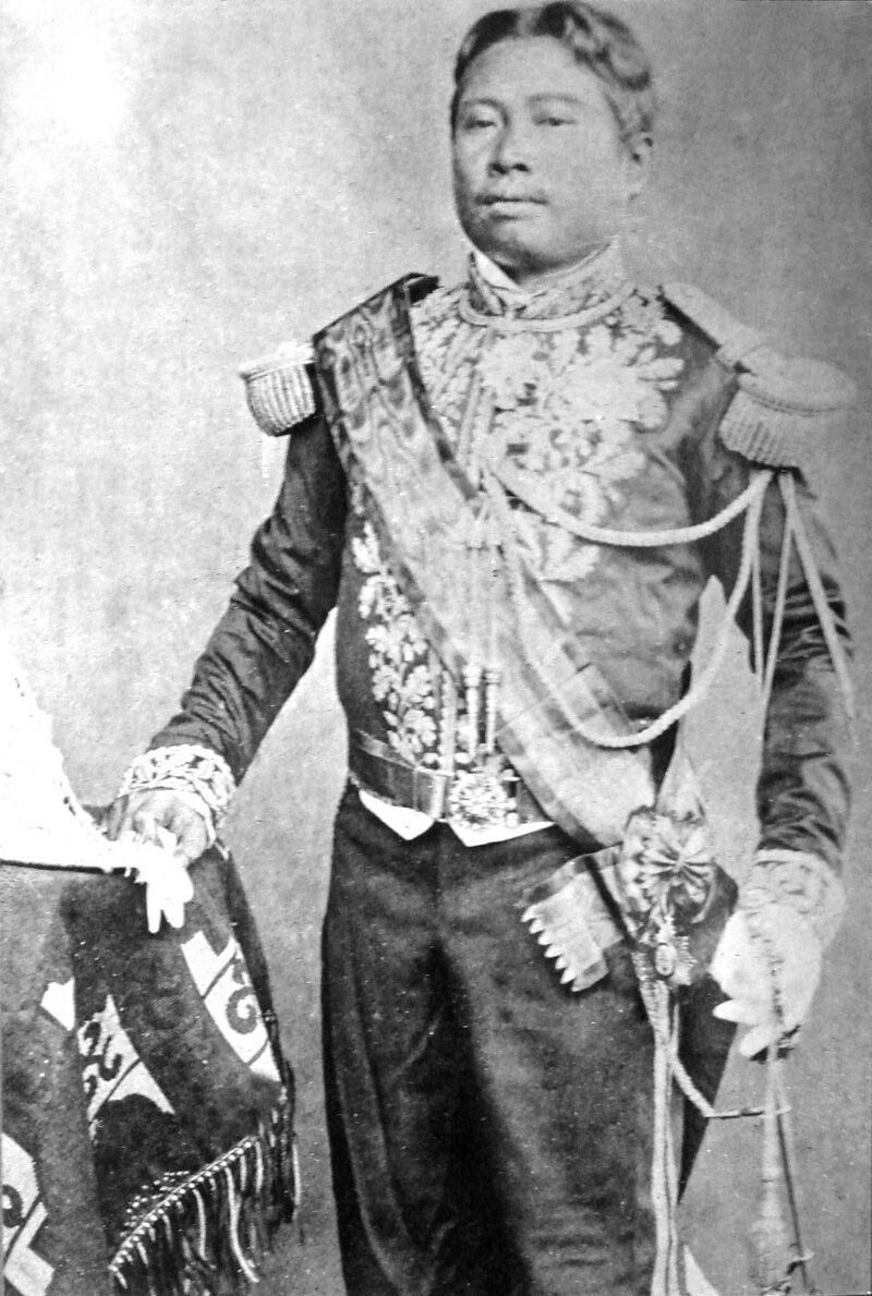 A black and white studio portrait of a standing man in an embroidered uniform jacket with epaulettes, a sash and medals, one hand resting on a draped table.