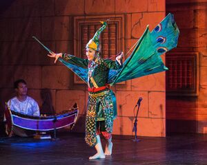 A dancer in a green jeweled costume and a tall peacock headdress spreads blue and green fabric wings painted with eyespots, stepping barefoot across a stage. A musician in white sits at a curved wooden xylophone on the left, in front of a painted stone temple backdrop.