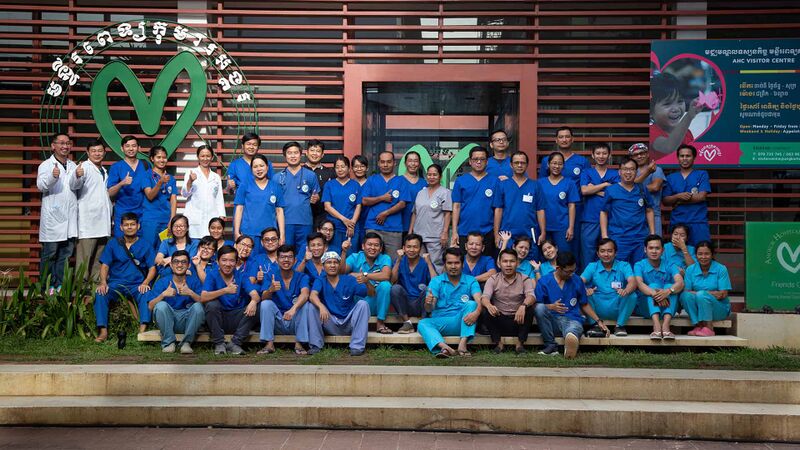 A large group of hospital staff in blue uniforms posed in front of a building with the hospital's green heart logo.