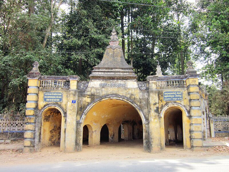 An arched stucco gateway with three openings, a tiered spire on top and tall trees behind.