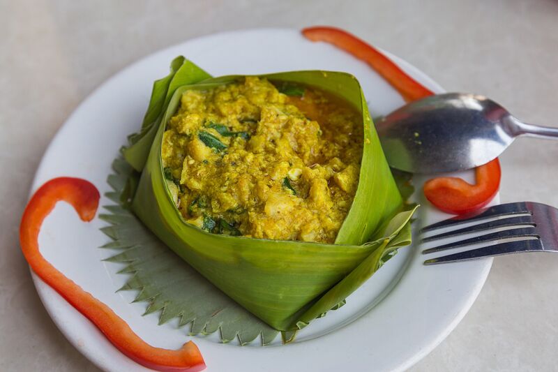 A square banana-leaf cup filled with yellow steamed fish curry on a white plate, garnished with red chilli.
