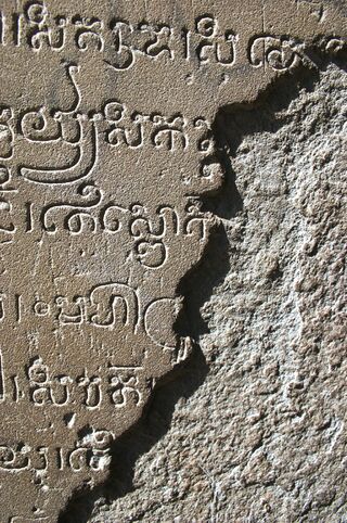 Close view of Khmer letters carved into a weathered stone surface, with a broken edge and trees in the background