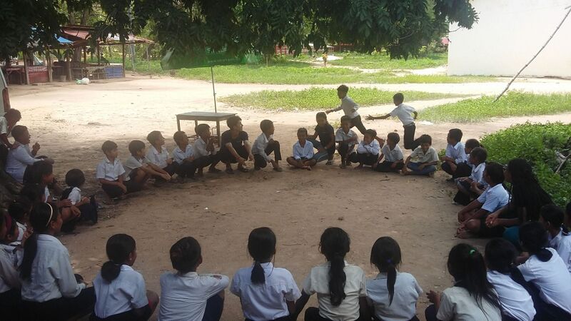 Dozens of uniformed students sit in a large circle on a dirt schoolyard under trees, seen from behind and at a distance.