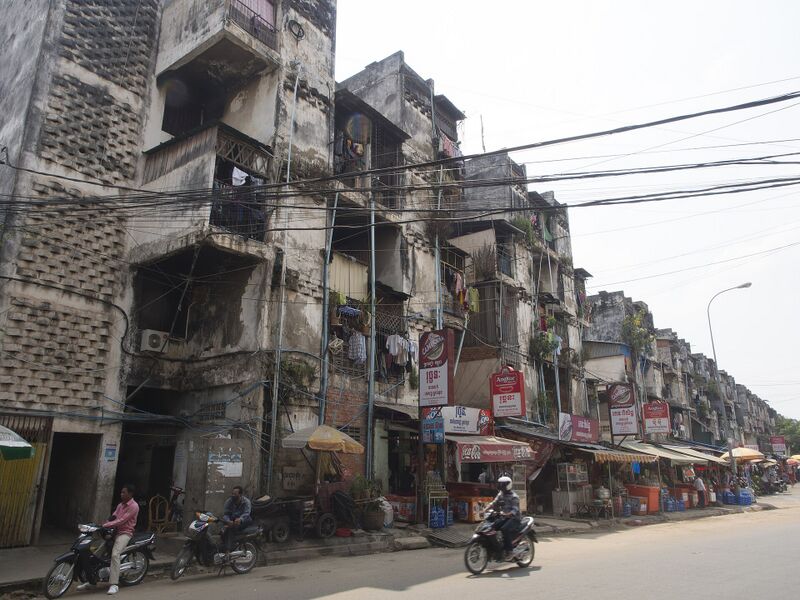 A long weathered apartment block above a busy street with shops and motorbikes.