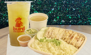 A tray of sliced poached chicken over rice with cucumber, small cups of sauce and soup, and a lemon drink.