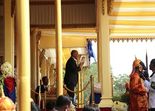 The king in a dark suit at a golden pavilion, with officials and a figure in red robes nearby.
